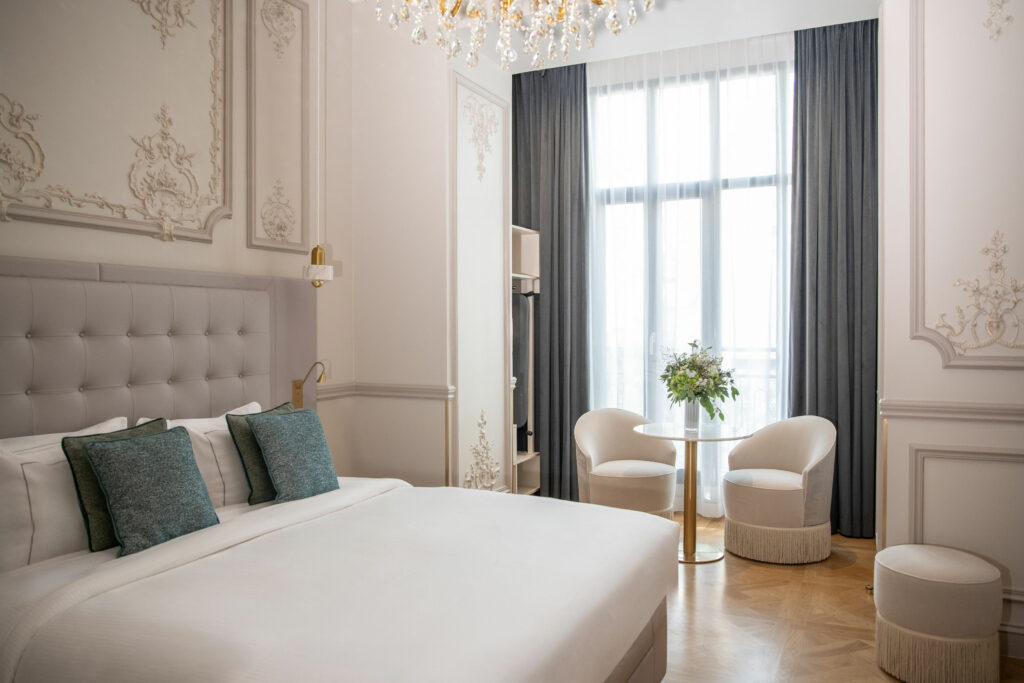 Chambre de luxe à Maison Triomphe Paris : coin salon avec fauteuils à franges, hautes fenêtres haussmanniennes, lustre en cristal et parquet en chêne, hôtel-résidence près de l’Arc de Triomphe